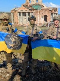 Vojáci drží vlajky před zničenými budovami v ukrajinské Klišivce
