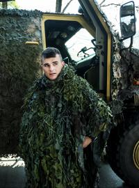 Voják oblékající si maskovací oděv hejkal před lehkým obrněným vozidlem Dingo