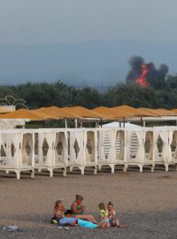 Lidé odpočívají na pláži, zatímco po explozích na ruské vojenské letecké základně v Novofedorivce na Krymu stoupá kouř a jsou vidět plameny, 9. srpna 2022